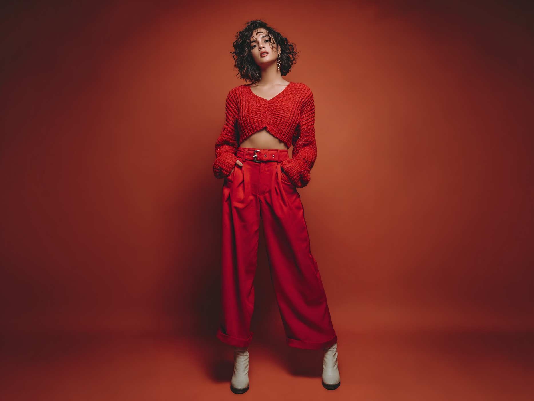 Eine Frau tr&auml;gt eine rote Stoffhose mit rotem Nieteng&uuml;rtel, dazu ein rotes, kurzes Stricktop und wei&szlig;e Boots.