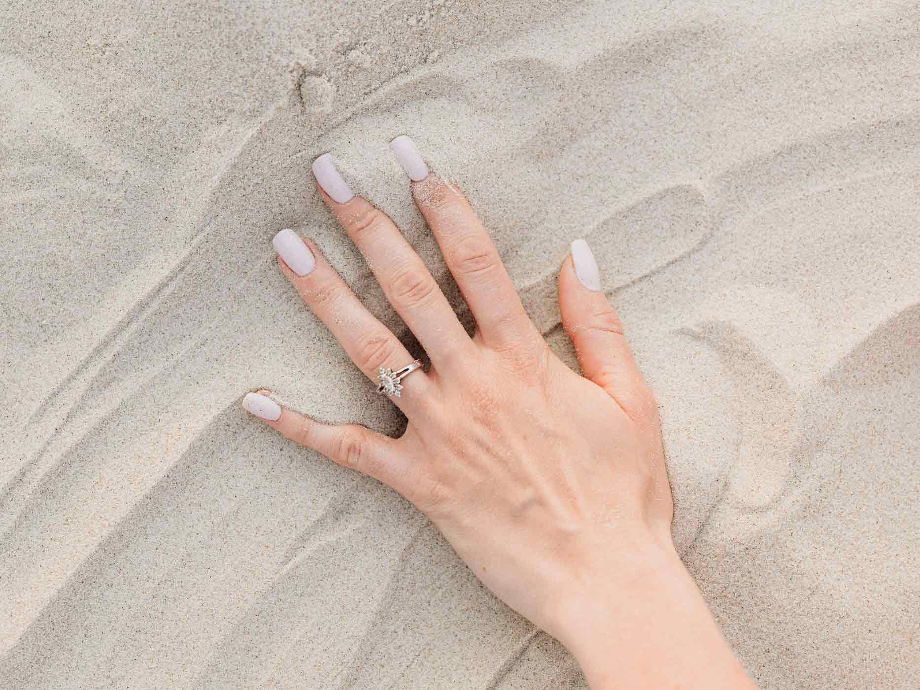 Eine Hand liegt auf Sand auf, die Fingernägel sind in einem zarten beigefarbenen Ton lackiert.