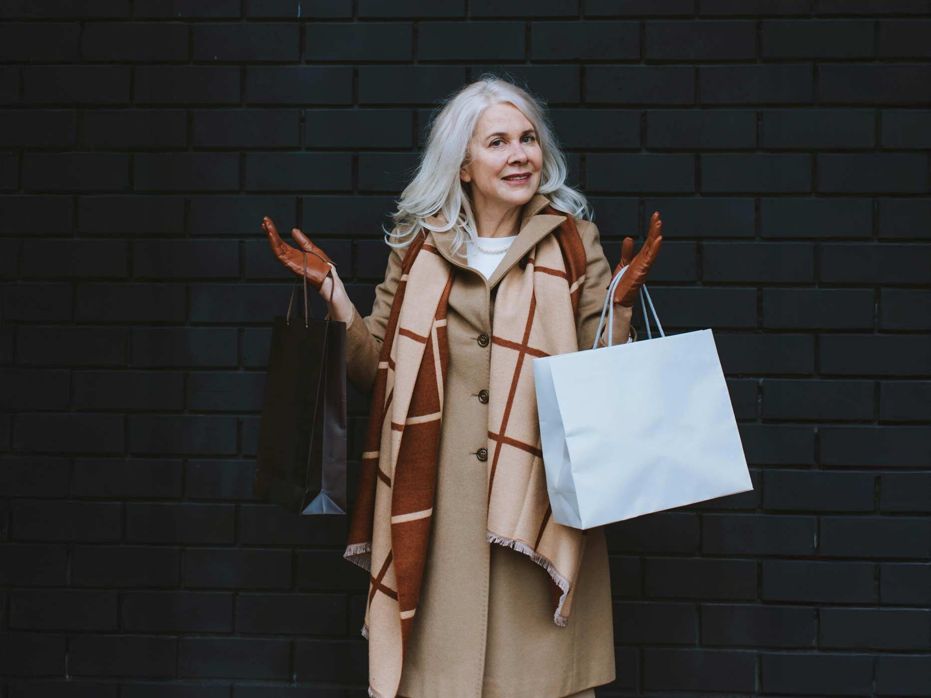 Eine Frau mit schulterlangen grauen Haaren tr&auml;gt einen langen beigen Mantel, dar&uuml;ber einen braunen Schal und braune Lederhandschuhe, sie h&auml;lt mehrere Einkaufst&uuml;ten in der Hand und zuckt mit den Schultern.