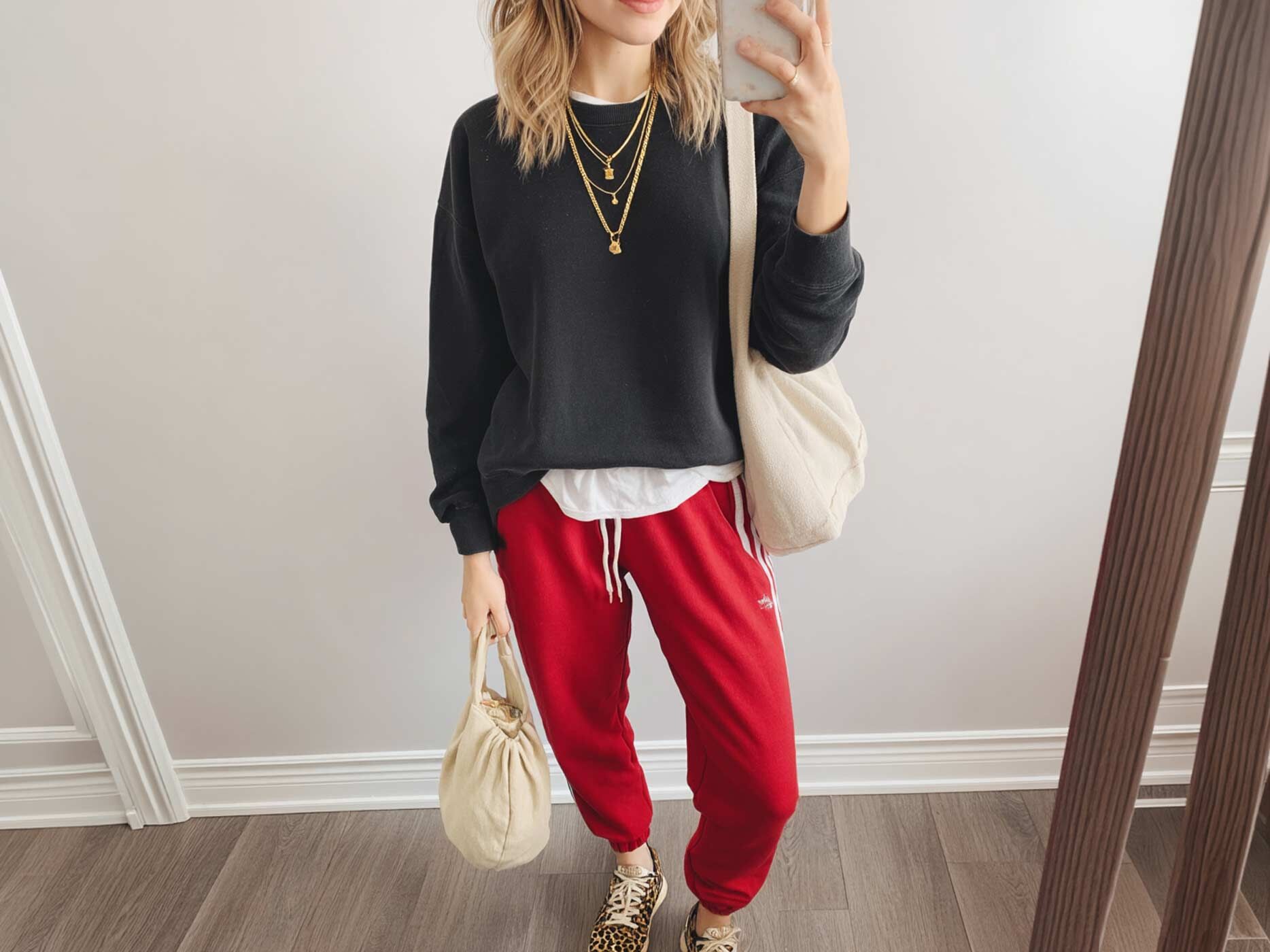 Eine Frau steht vor einem Spiegel, sie tr&auml;gt eine rote Jogginghose, ein wei&szlig;es Hemd, dar&uuml;ber einen schwarzen Pullover und goldenen Schmuck. Sie h&auml;lt zwei wei&szlig;e Taschen in der Hand und ihre Schuhe zieren ein Leo-Muster.