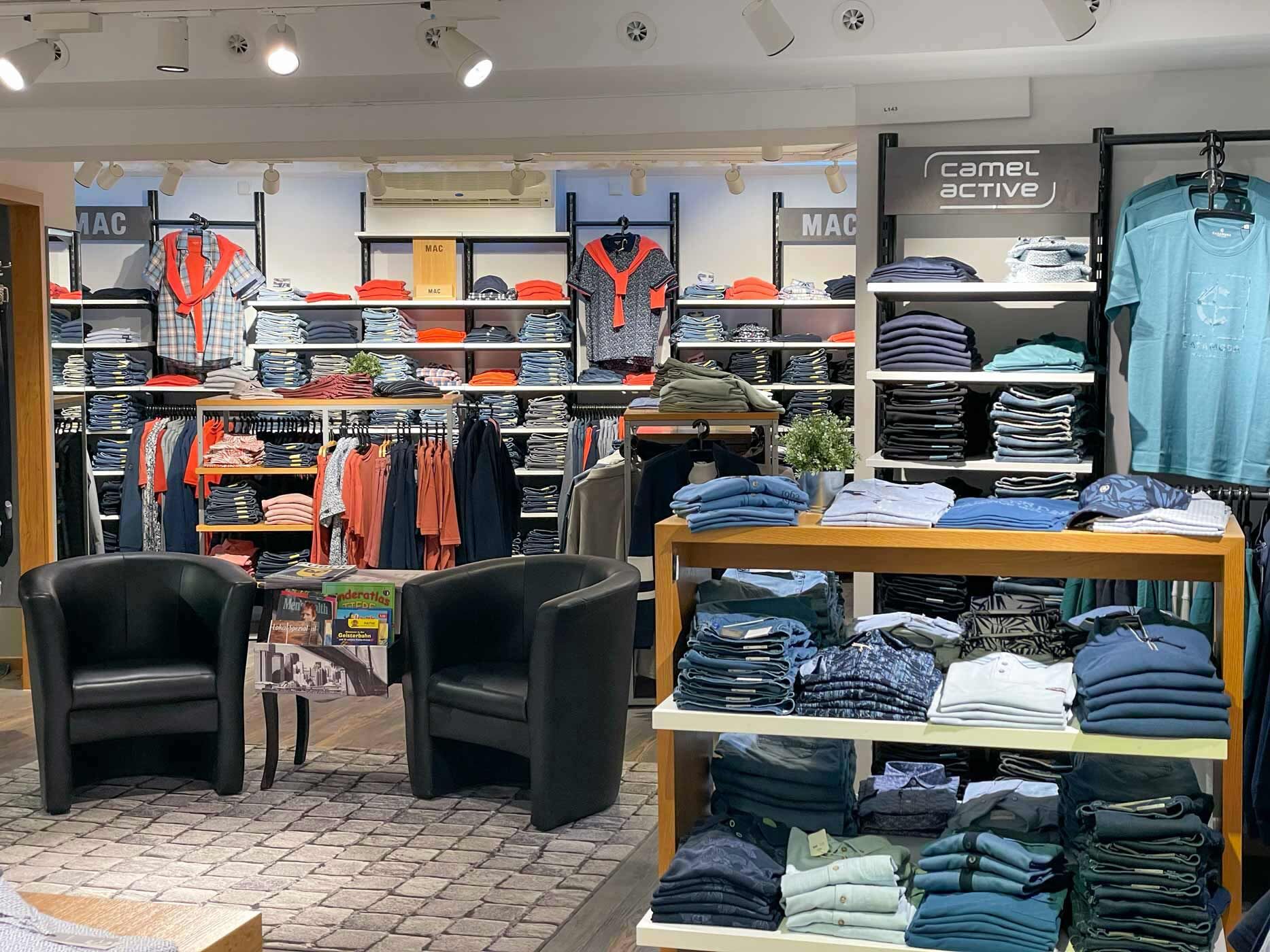 Heller Verkaufsraum eines Modegesch&auml;fts mit ordentlich pr&auml;sentierten Jeans, Shirts und Oberteilen verschiedener Marken. Zwischen den Regalen stehen zwei schwarze Sessel, die zum Verweilen und zur Beratung einladen.