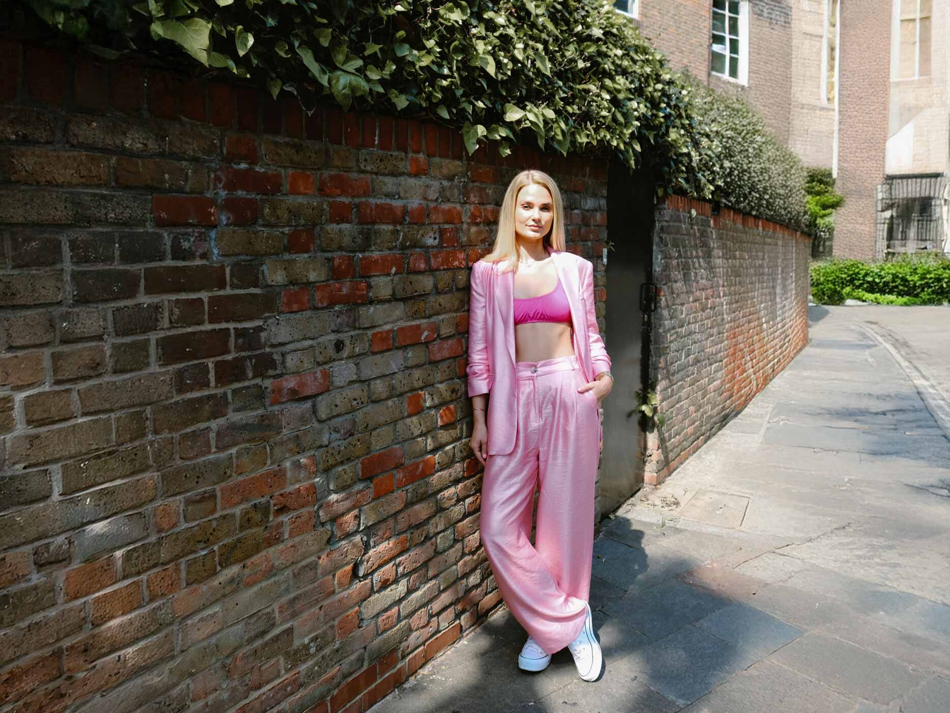 Eine blonde Frau lehnt an einer Mauer, sie trägt eine weite, rosa Stoffhose, einen knallpinken BH und einen rosa Blazer.