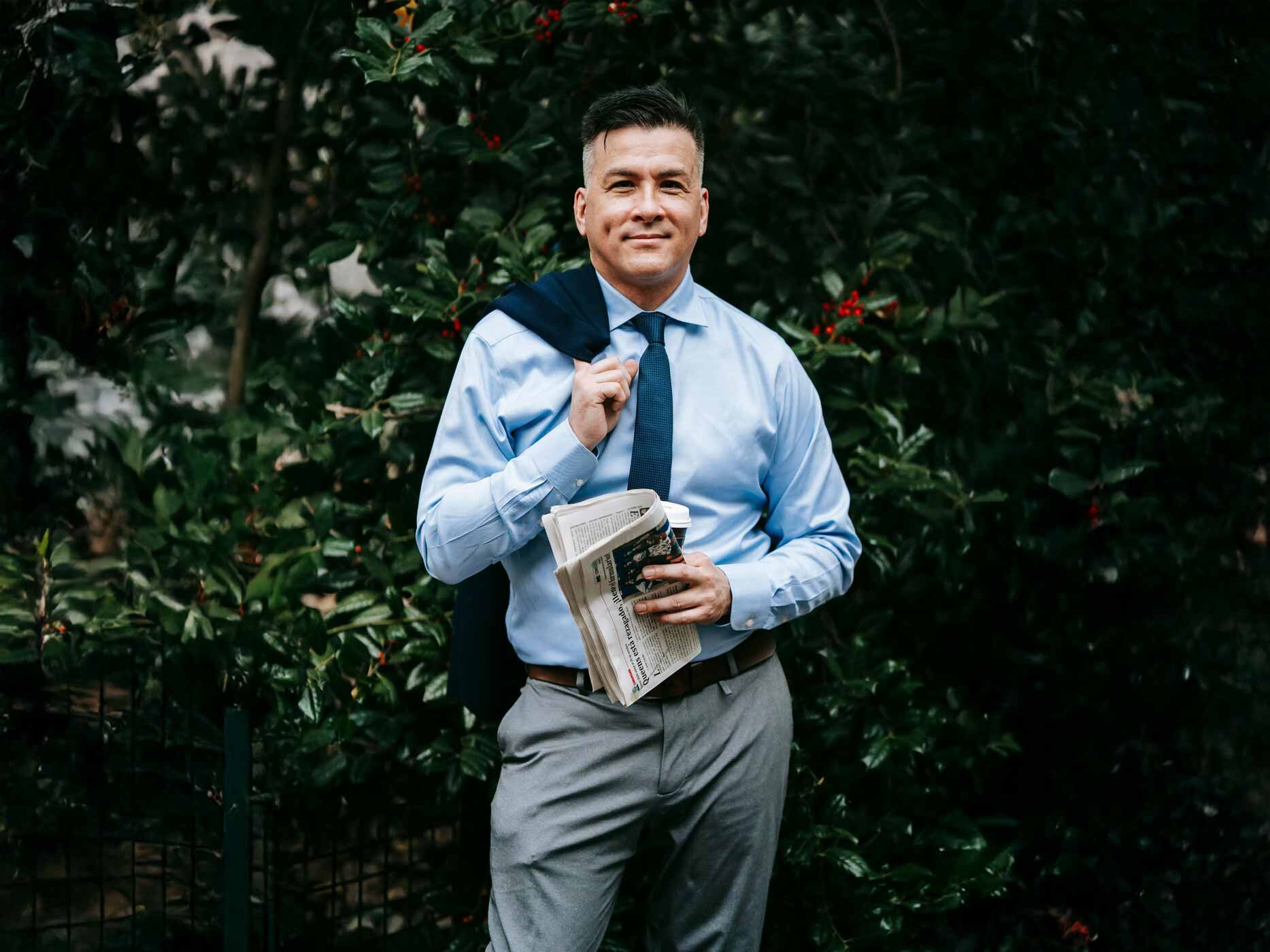 Ein Mann trägt eine graue Anzugshose, ein hellblaues Hemd und eine blaue Krawatte, er hat eine Zeitung in der Hand und posiert für die Kamera.