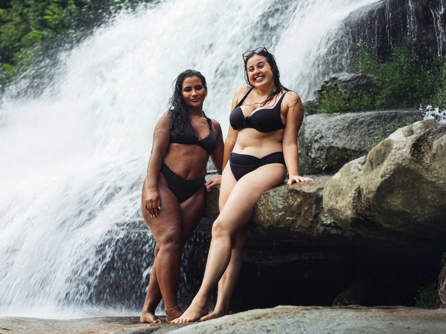 Zwei kurvigere Frauen tragen schwarze Bikinis und posieren vor einem Wasserfall.