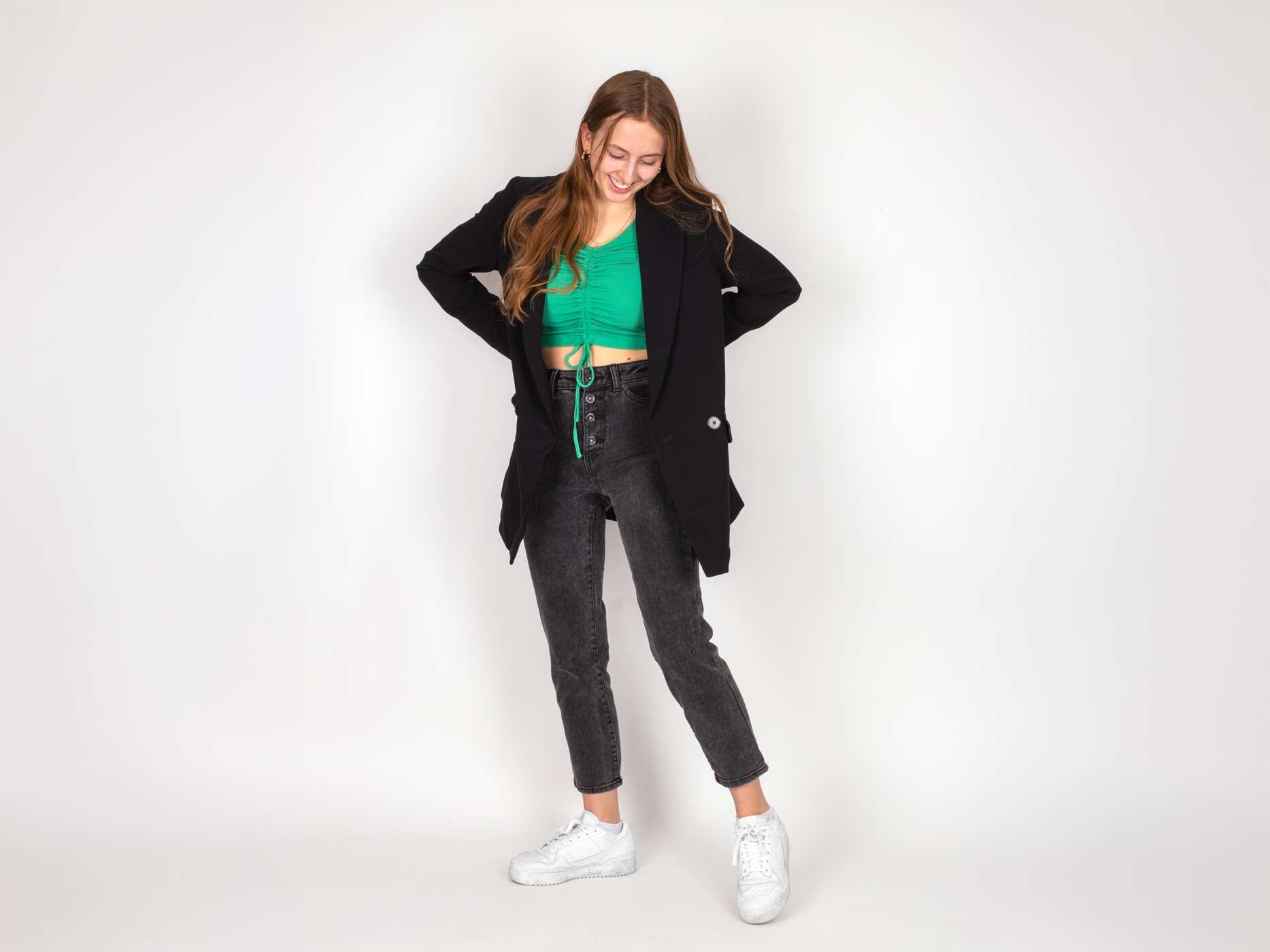 Ein Mädchen trägt eine graue Hose, dazu ein grünes Top, einen schwarzen Blazer und weiße Sneaker.