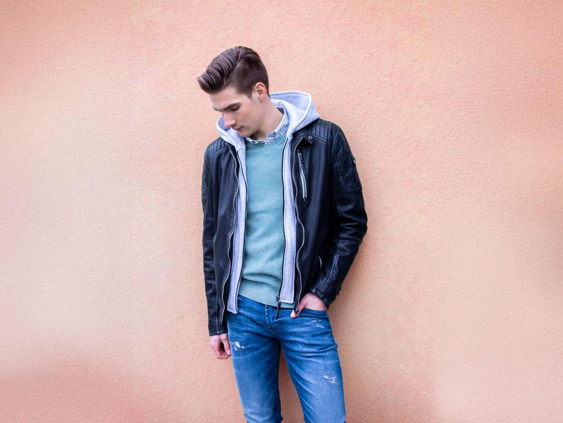 Ein junger Mann lehnt an einer Wand, er trägt eine blaue Jeans, ein blauen Pullover und darüber eine graue Stoffjacke und eine schwarze Lederjacke.