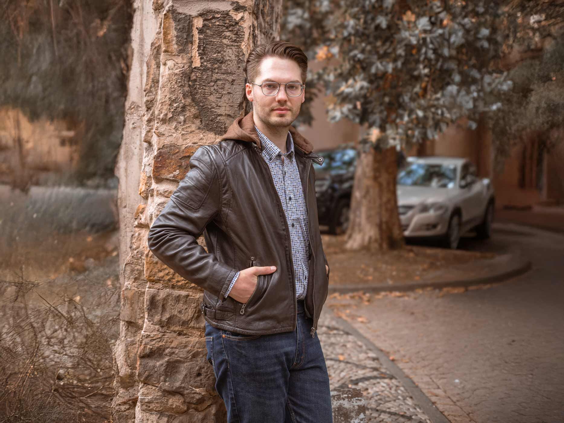 Ein junger Mann lehnt an einem Baum, er trägt ein weißes kariertes Hemd und eine braune Lederjacke.