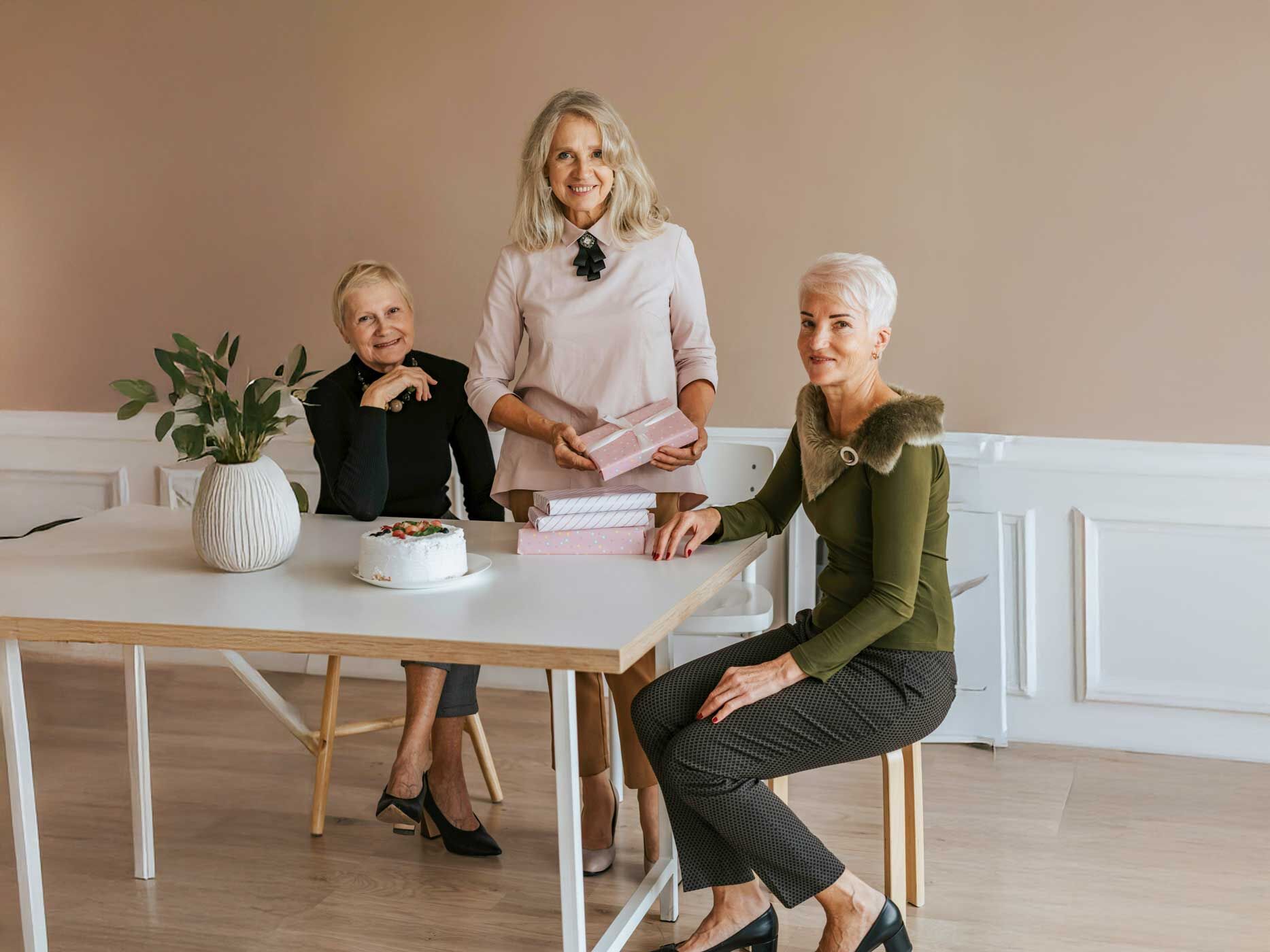 Drei ältere Frauen tragen schlichte, elegante Kleidung in ruhigen Farben wie Schwarz, Rosé, Beige und Olivgrün mit klaren Schnitten und hochwertigen Stoffen. Die Outfits wirken klassisch und modern zugleich, mit Blusen, feinen Stoffhosen und Pumps, abgestimmt auf graues und weißes Haar.