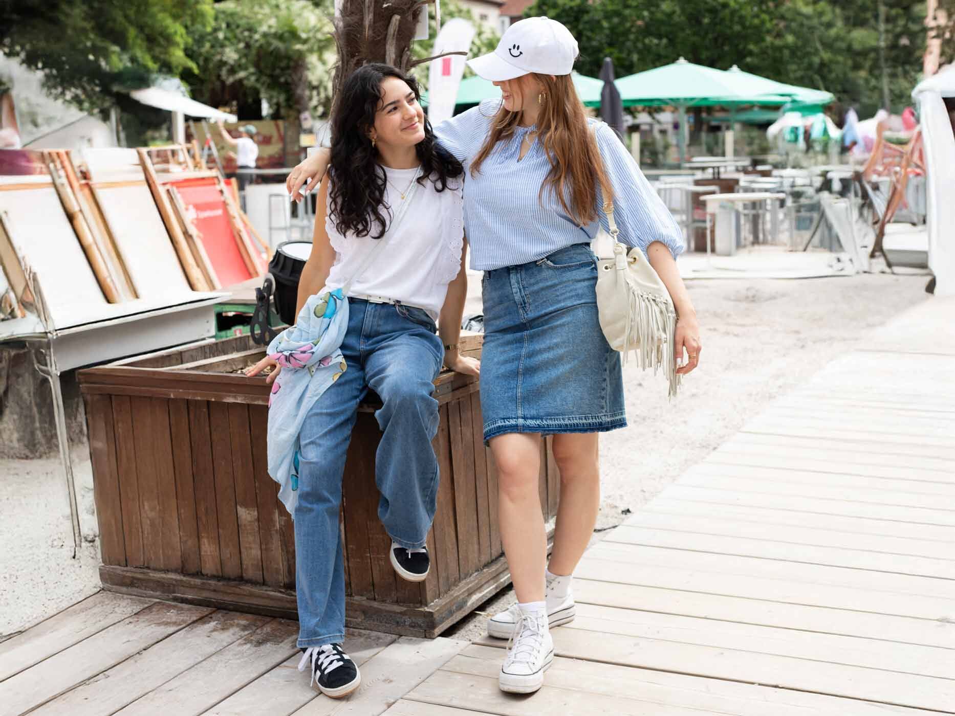Zwei junge Mädchen sitzen an einer Palme des Citybeachs. Sie tragen moderen Outfits mit Denimstoff.