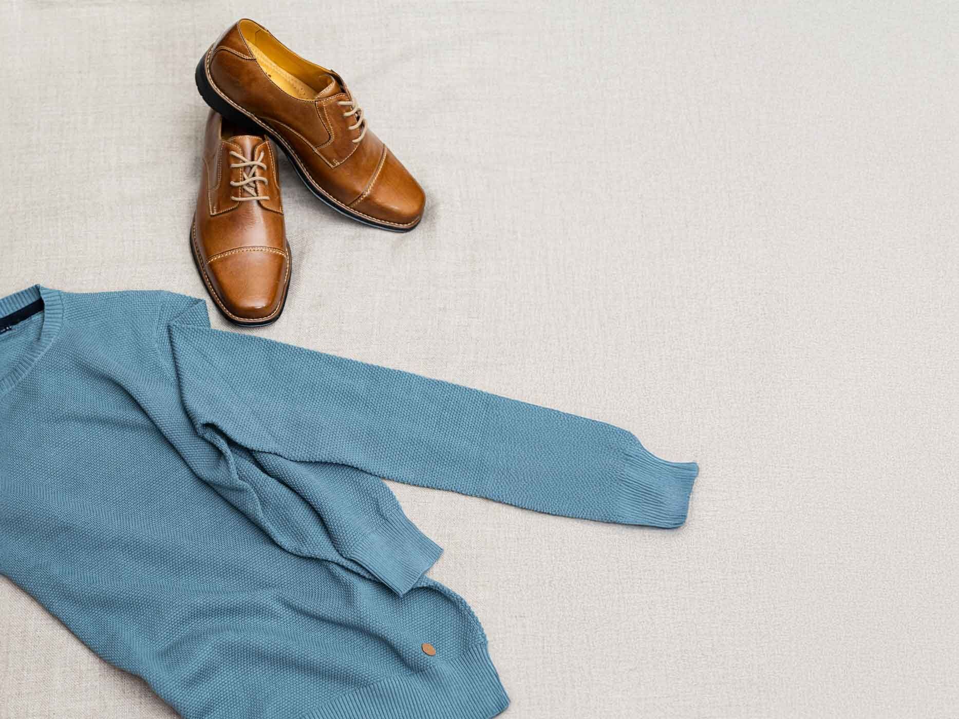 Ein blauer Pullover liegt gefaltet auf einem weißen Teppich, daneben liegt ein Paar braune Herrenschuhe.