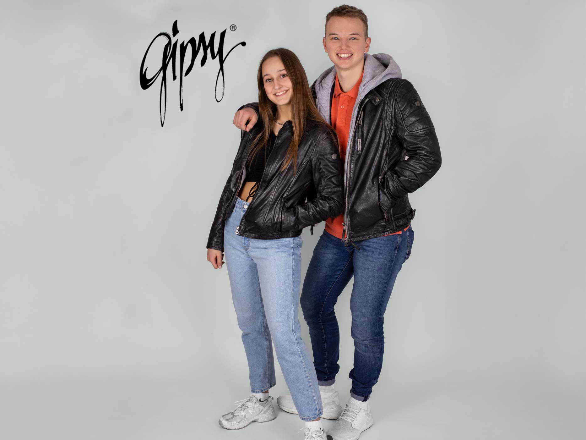 Ein junge legt seinen Arm und ein Mädchen, sie tragen beide blaue Jeans und eine schwarze Lederjacke und lächeln in die Kamera.