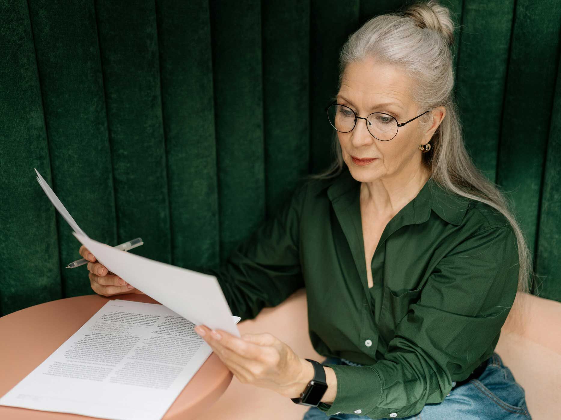 Eine Frau mit langen grauen Haaren sitzt an einem kleinen Tisch, sie trägt eine tief dunkelgrüne Bluse und eine blaue Hose, dazu eine Brille.