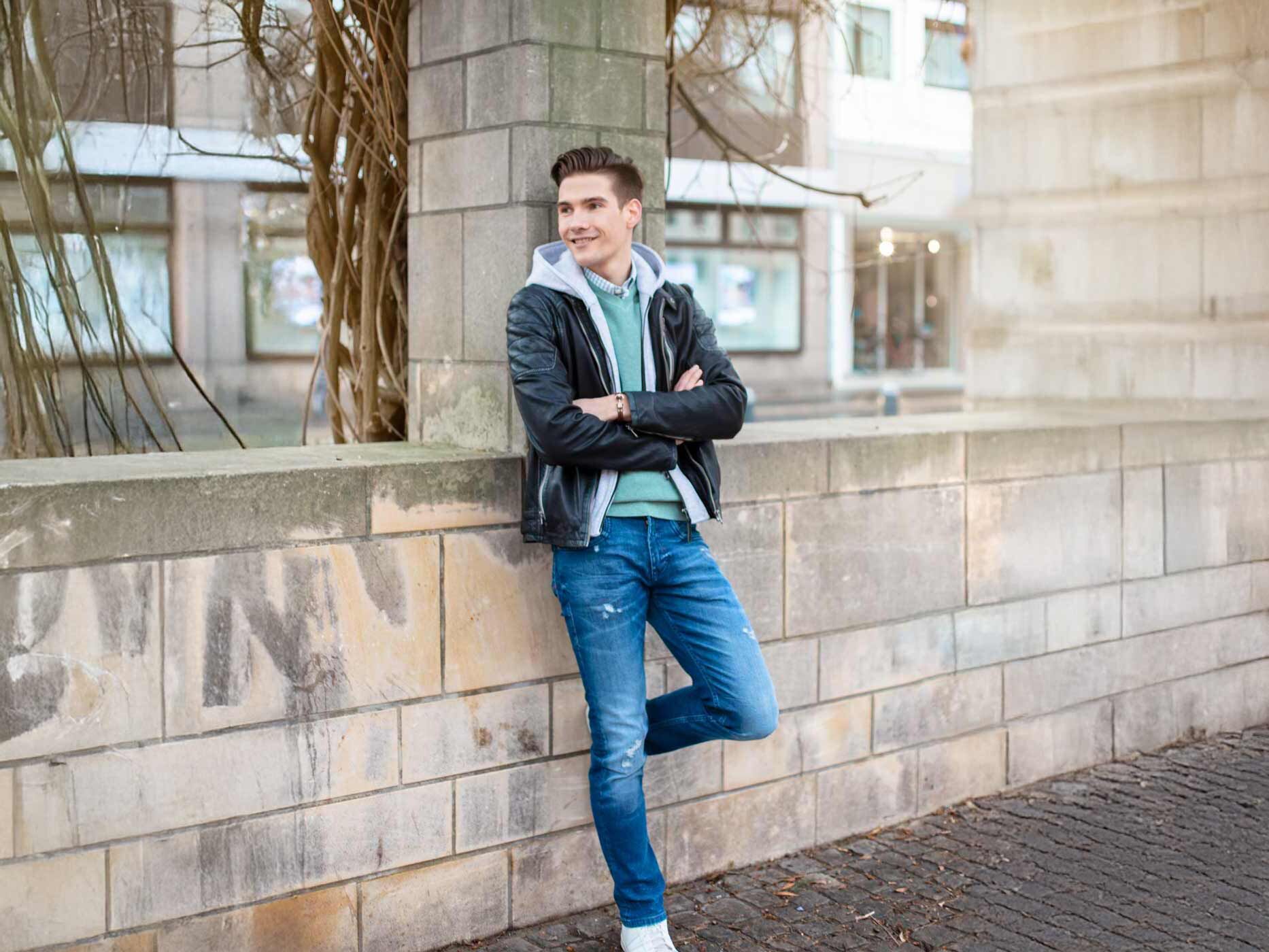 Ein Mann lehnt an einer Mauer und trägt eine blaue Jeans, dazu einen blauen Pullover, eine graue Stoffjacke und eine schwarze Lederjacke.