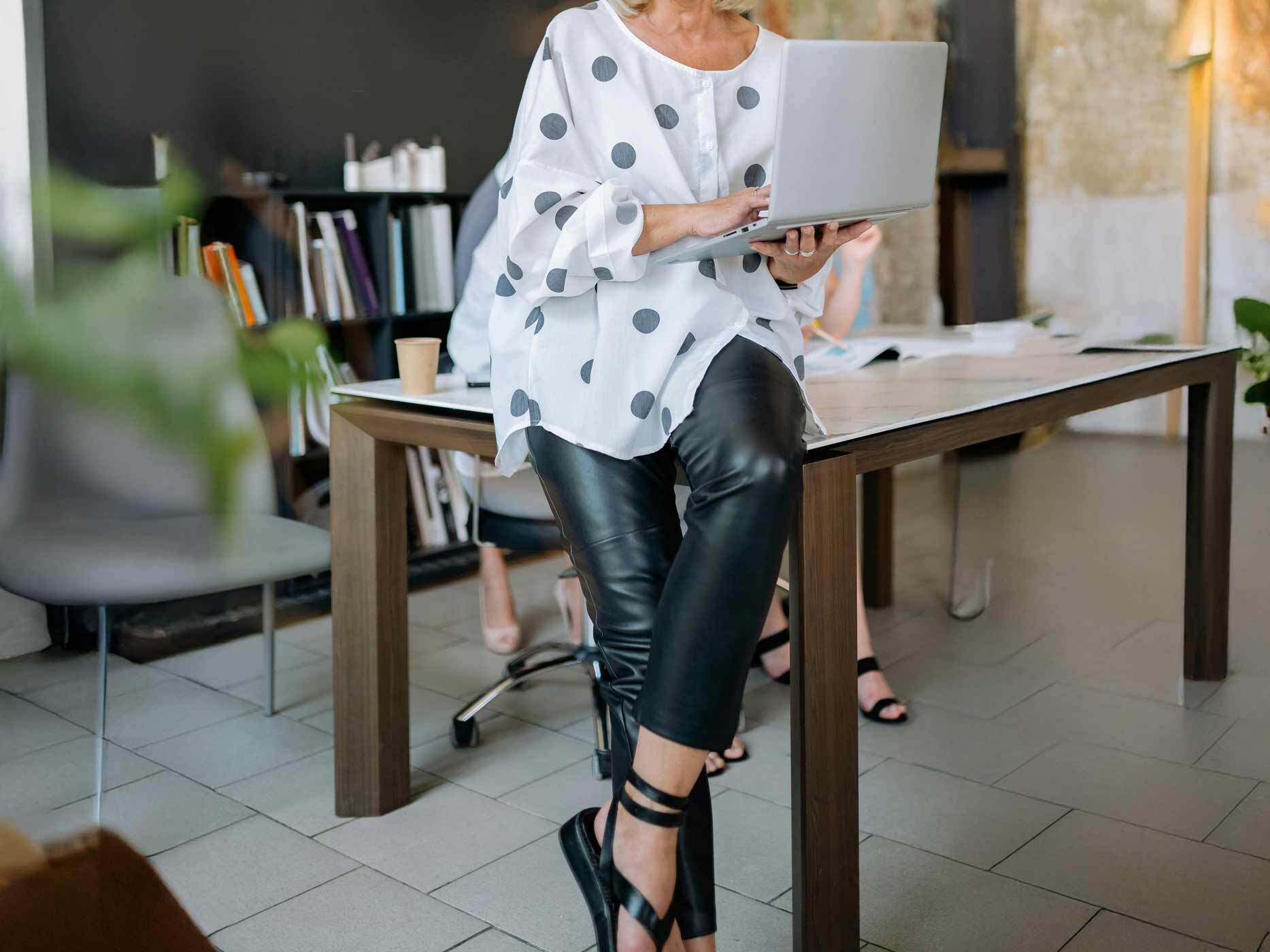 Frau in schwarzer Lederhose und mit Oversize-Bluse sitzt mit überschlagenen Beinen und einem Laptop in der Hand auf der Tischkante.