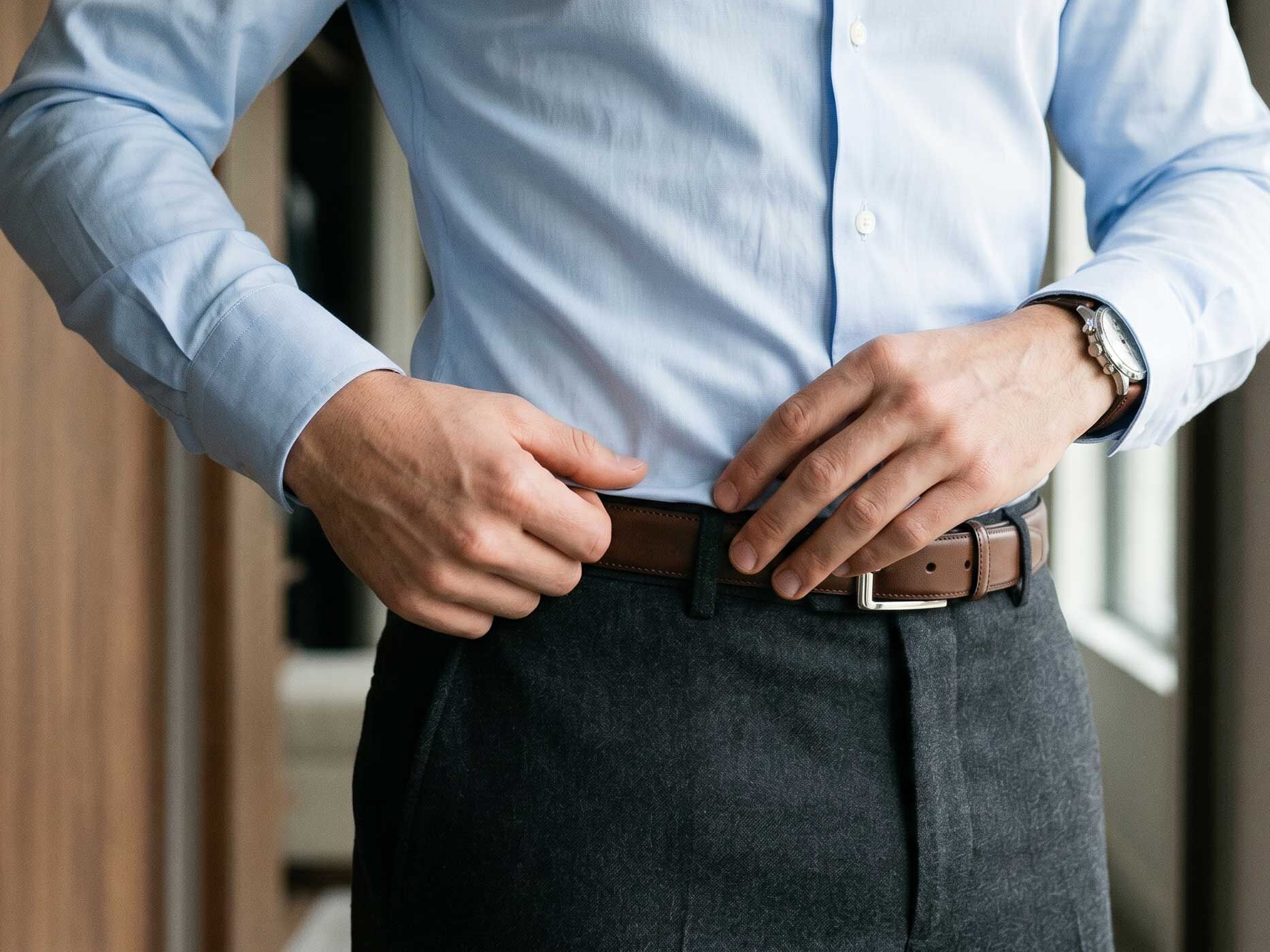 Nahaufnahme eines Mannes, der ein hellblaues Herrenhemd ordentlich in eine dunkle Stoffhose steckt und dabei seinen braunen G&uuml;rtel zurechtr&uuml;ckt.