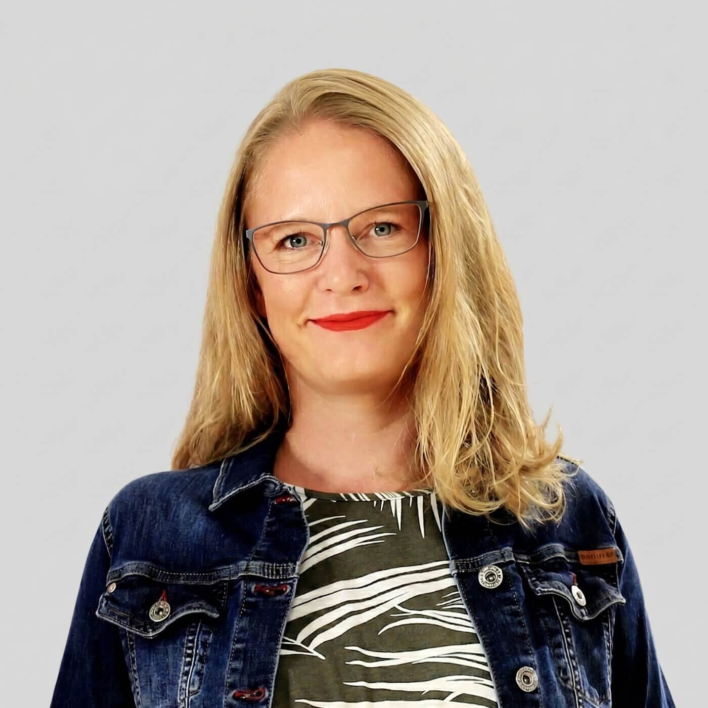 Aren Portraitfoto von Modeexpertin Ina. Sie hat blonde Haare, trägt eine Brille und lacht.