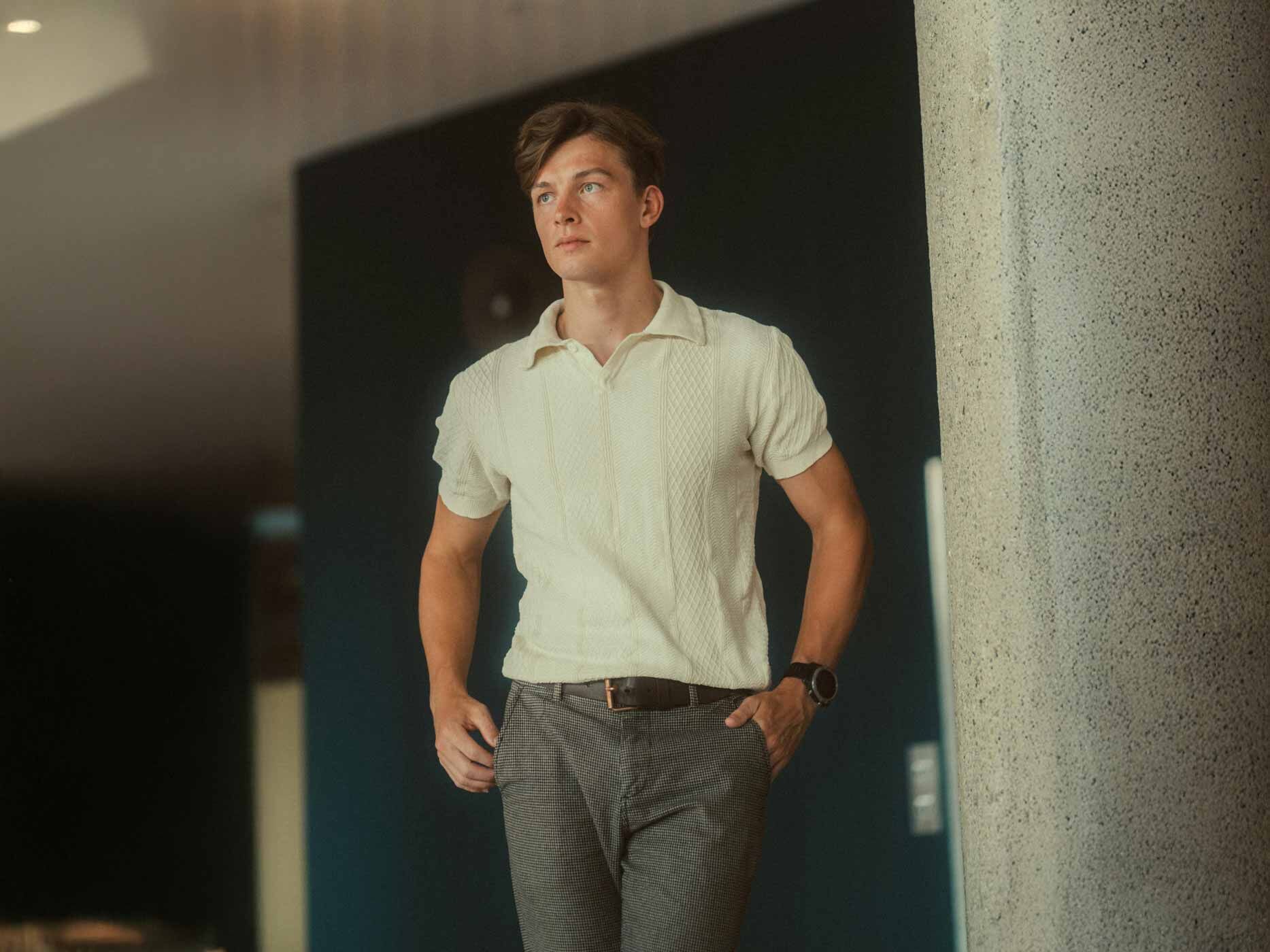 Ein junger Mann trägt eine graue Stoffhose, einen dunklen Gürtel und ein weißes Poloshirt in Strickoptik.