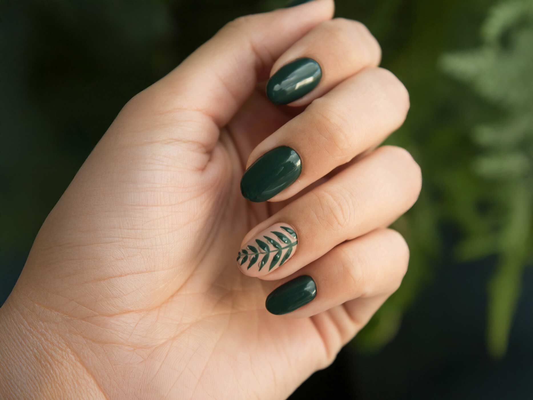 Fingernägel, die bis auf einen alle dunkelgrün lackiert sind. Auf dem einen Fingernagel ist ein Pflanzen-Motiv lackiert.