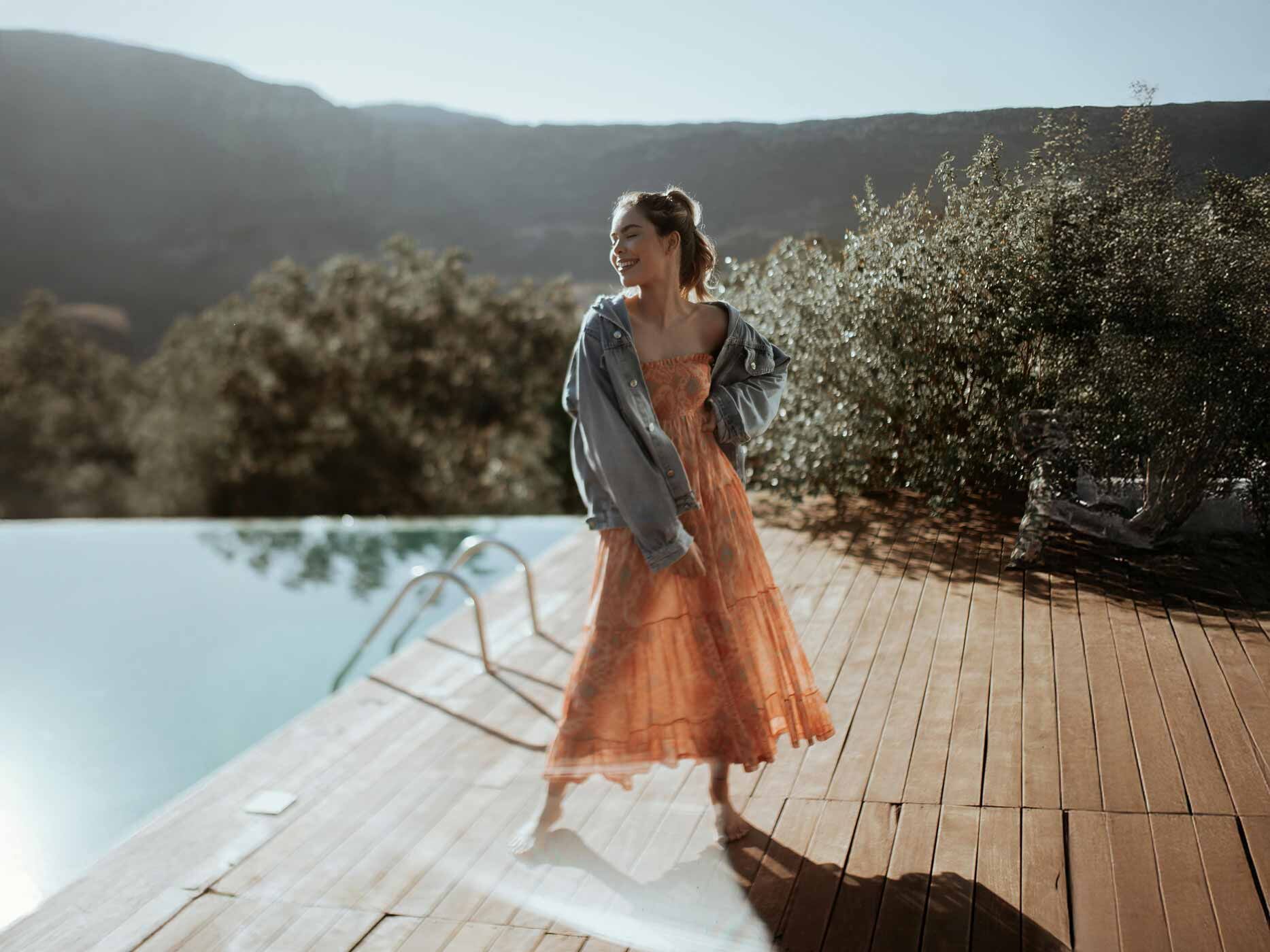 Eine Frau steht in der Sonne an einem Pool, sie trägt ein langes, luftiges orangenes Kleid und dazu eine locker geschnittene blaue Jeansjacke.