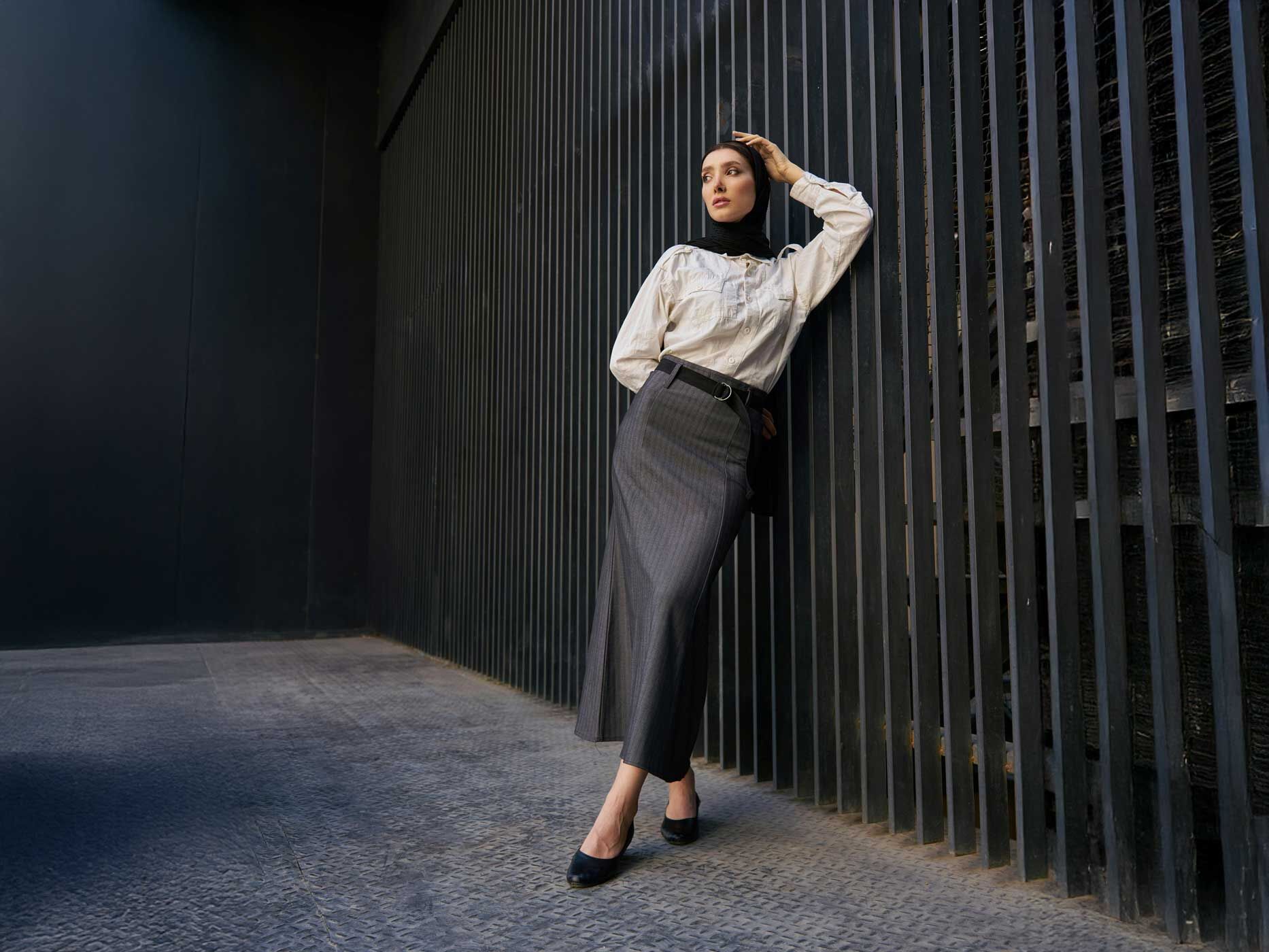 Eine Frau in einem langen Rock und mit Hijab, lehnt an einer Wand. Ihr Rock ist grau mit Streifen und ihre Bluse weiß.
