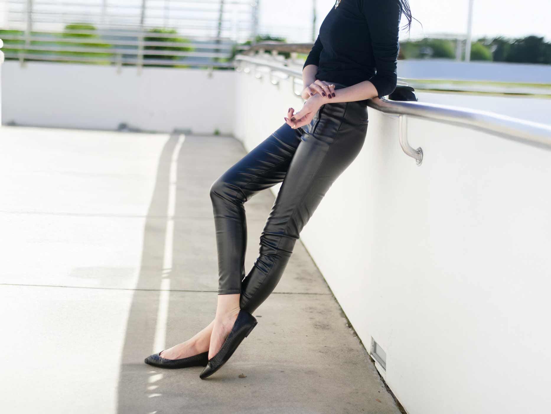 Frau in enger schwarzer Lederhose und Ballerinas lehnt an einem Geländer von einem Parkhaus oder Ähnlichem.