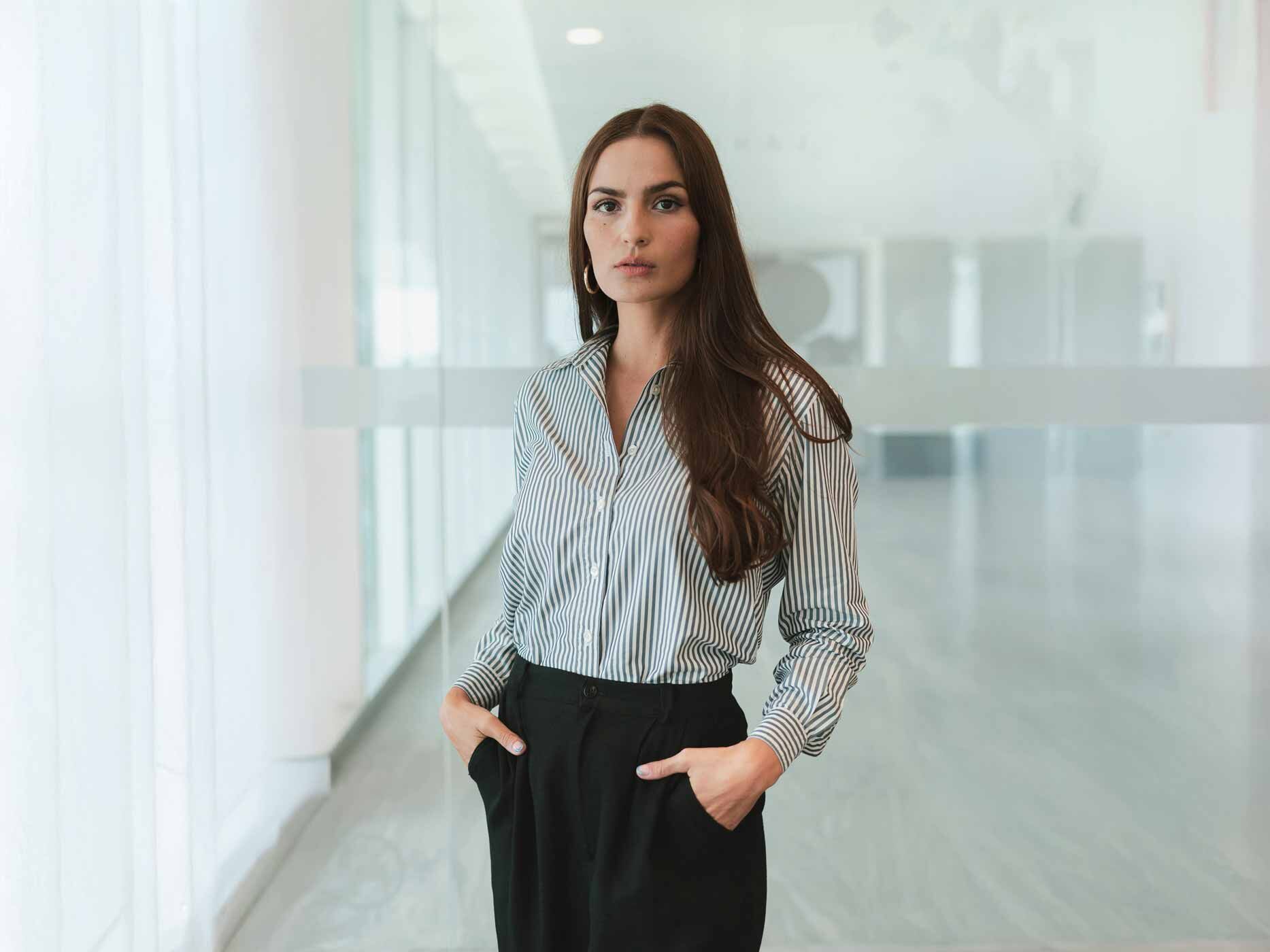 Eine Frau steht in einem B&uuml;roflur und tr&auml;gt eine schwarze Stoffhose und eine grau-wei&szlig; gestreifte Bluse.