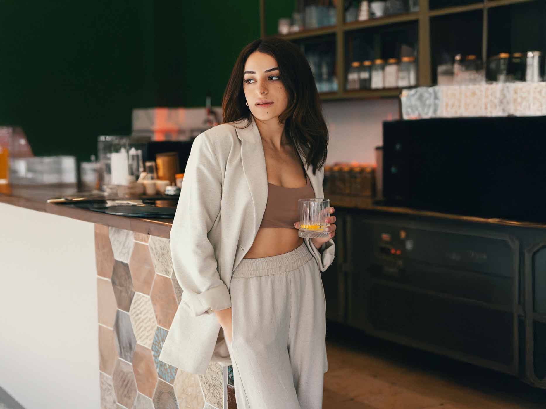Eine Frau steht an einer Bar und hat ein Glas in der Hand, sie trägt eine beige Jogginghose, einen beigen Blazer und ein kurzes, braunes Top.