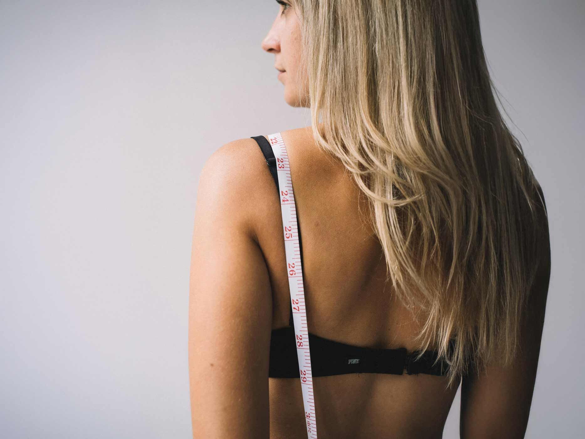 Eine Frau steht mit dem Rücken zur Kamera, trägt einen schwarzen BH und hat ein Maßband über der Schulter liegen.