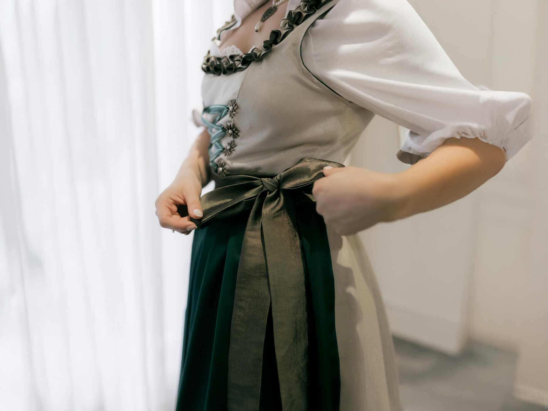 Eine Frau wird von der Seite gezeigt, sie bindet sich die Dirndl Schleife ihres dunkelgrünen Dirndls an der linken Seite.