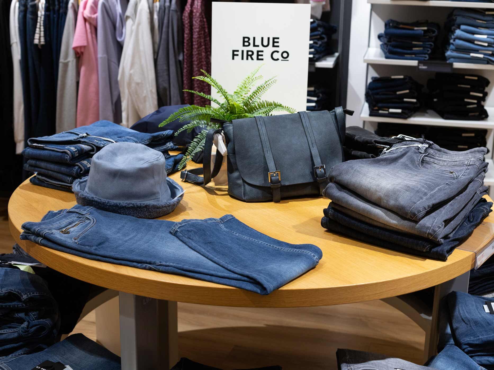 Auf einem runden Holztisch in einem Modegesch&auml;ft liegen mehrere gefaltete Damenjeans in verschiedenen Waschungen. Daneben stehen eine Handtasche, ein Hut und ein Schild der Marke Blue Fire, w&auml;hrend im Hintergrund weitere Kleidungsst&uuml;cke pr&auml;sentiert werden.