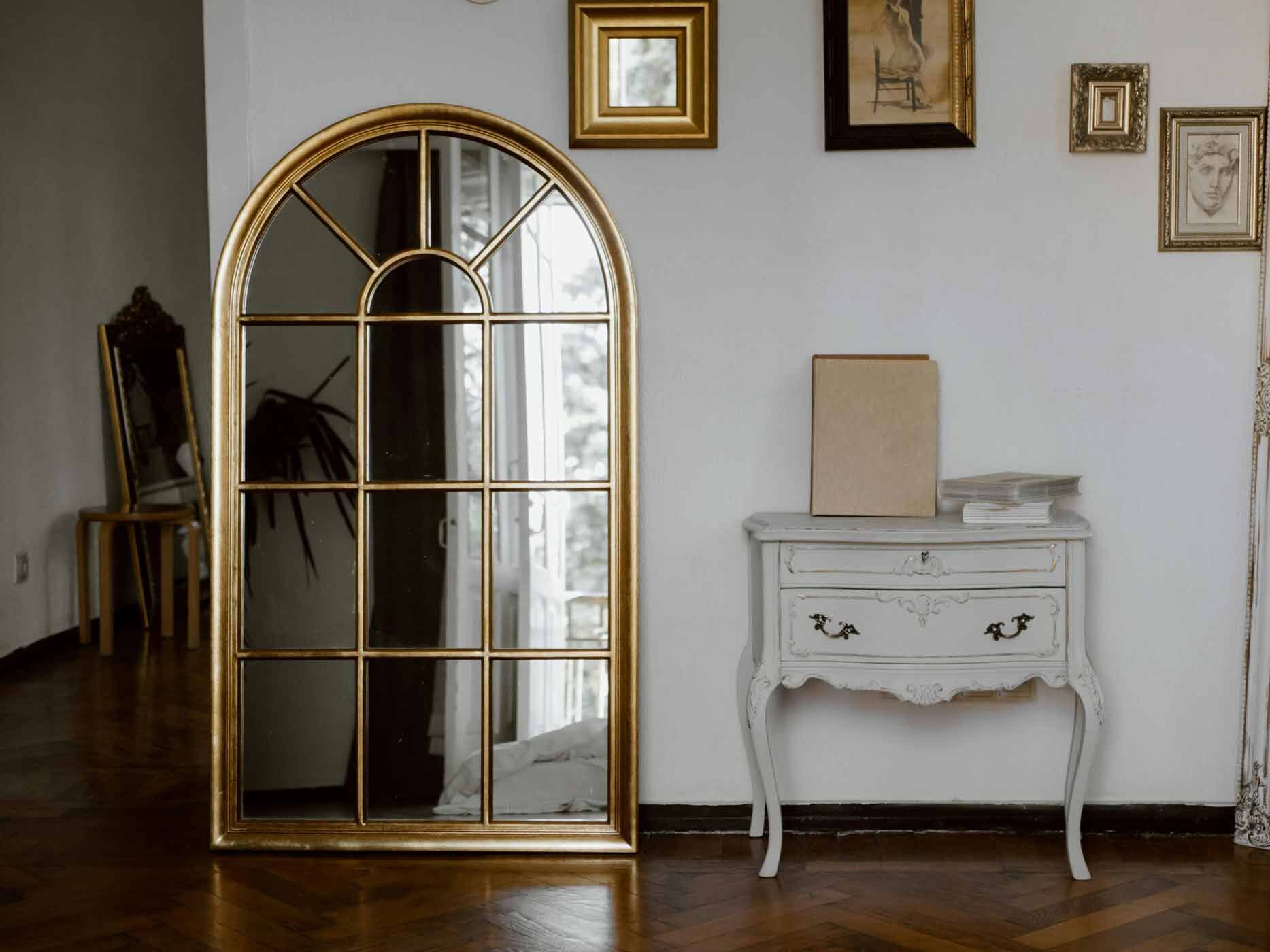 Ein Spiegel mit goldener Verzierung steht neben einer kleinen, weißen Kommode vor einer weißen Wand.