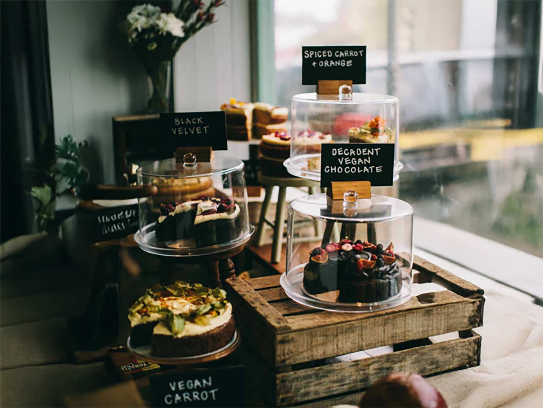 Mehrere Kuchen und Torten stehen unter Glashauben und mit Schildern ausgezeichnet in einem Fensterbereich eines Caf&eacute;s.