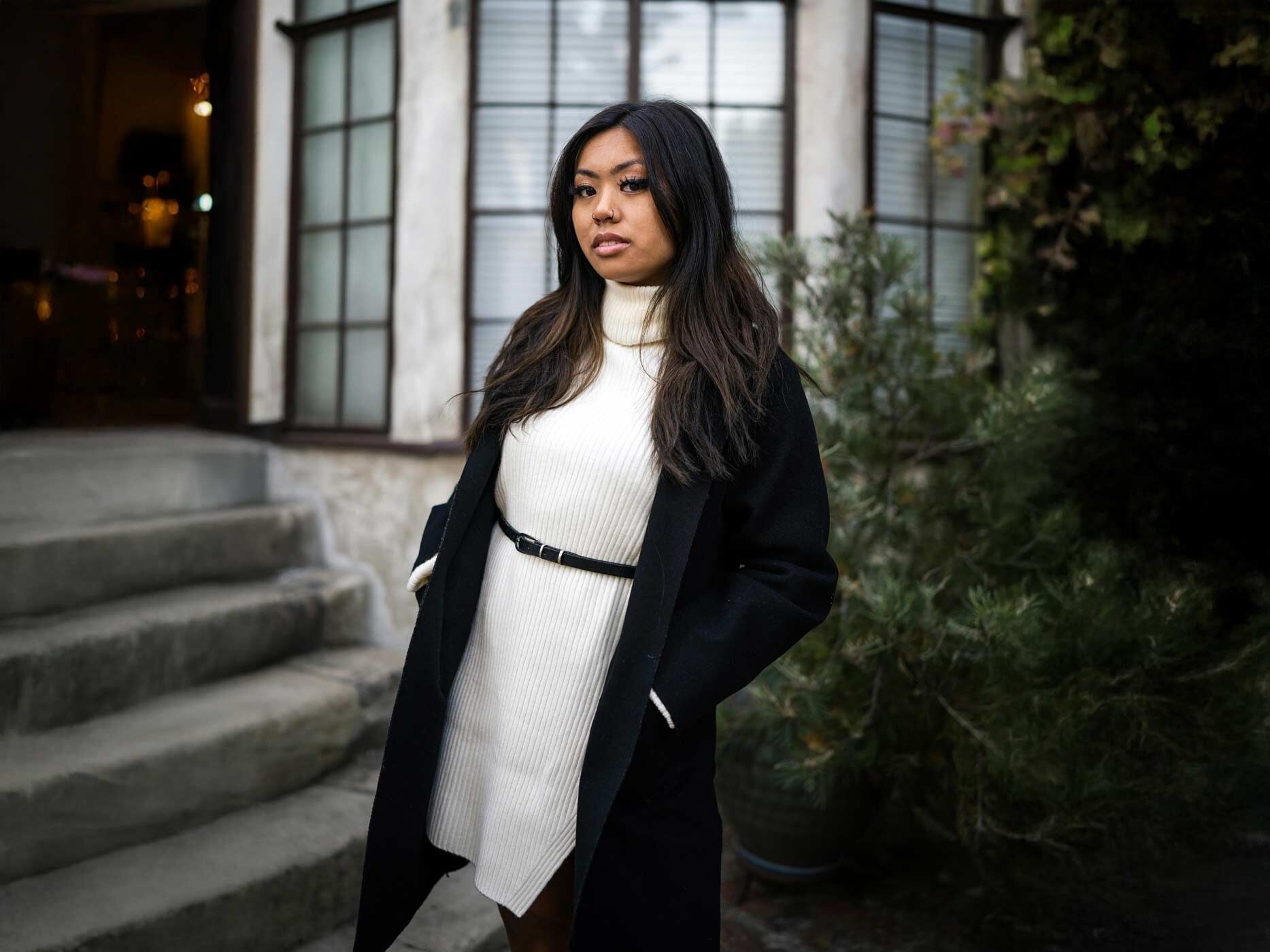 Eine Frau trägt ein weißes Strickkleid, dazu einen schwarzen Tailliengürtel und einen schwarzen Mantel.