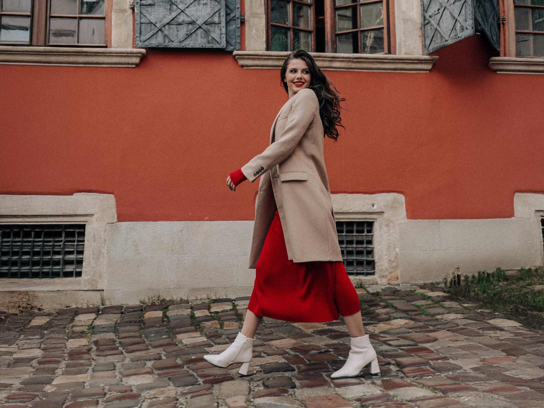 Eine Frau trägt ein langes rotes Kleid, darüber einen braunen Trenchcoat und weiße Stiefeletten, sie spaziert an Häusern entlang.