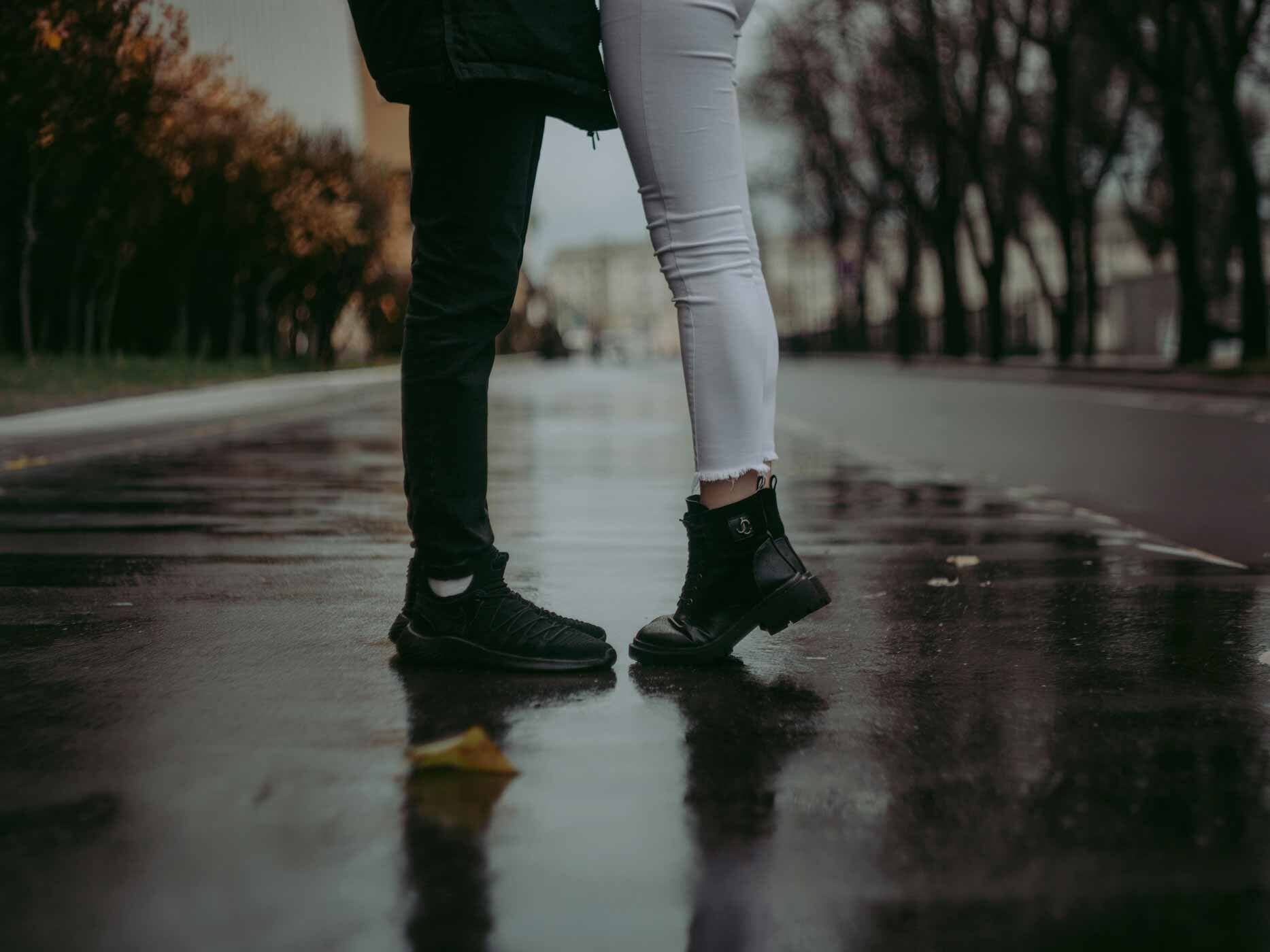 Zwei Personen stehen sich auf nassem Asphalt gegen&uuml;ber, zu sehen sind nur die Beine. Die Frau tr&auml;gt eine Kombination aus heller Jeans markante schwarzen Boots