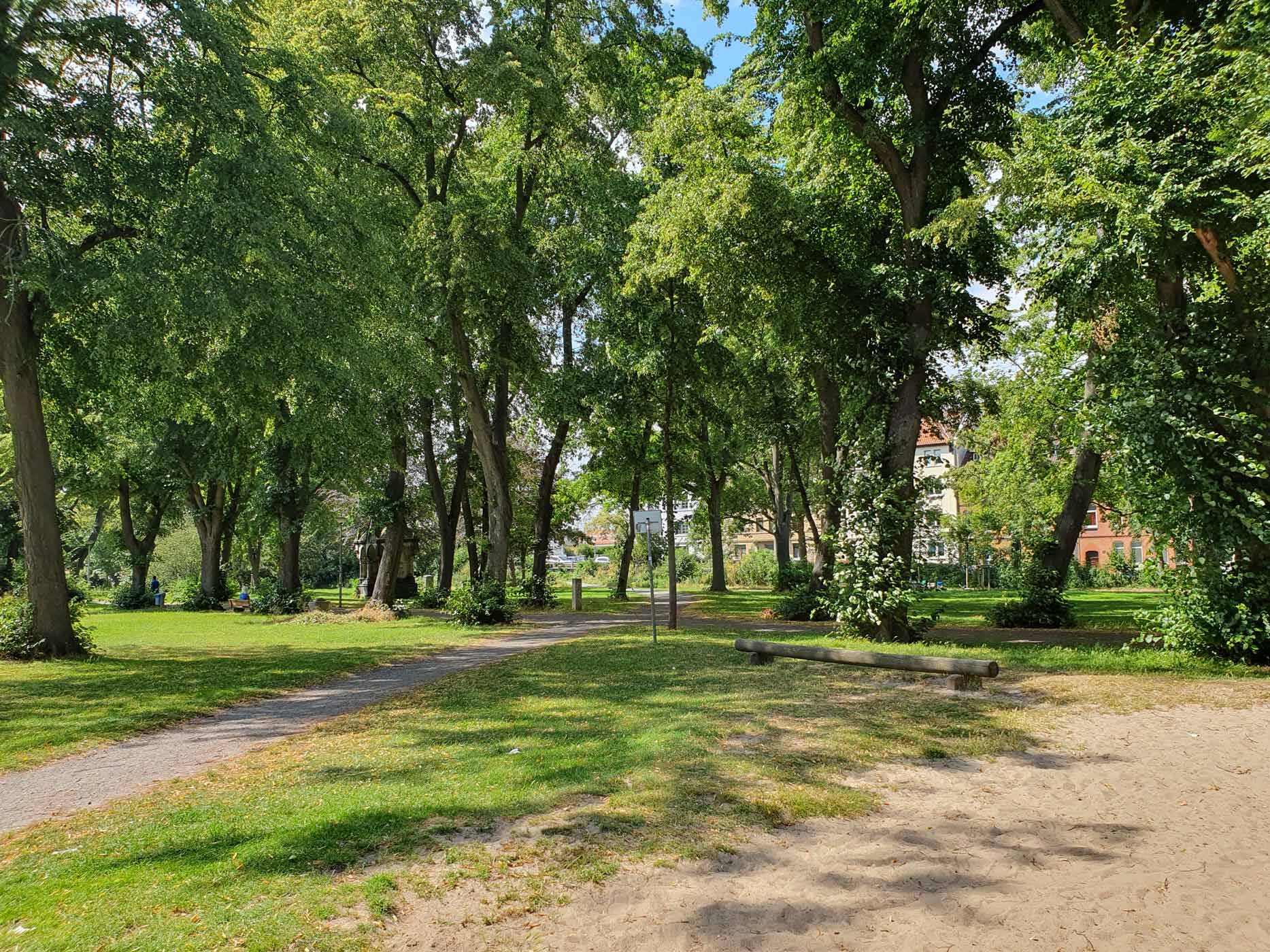 Grünfläche mit vielen Bäumen, einem Baumstamm zum Sitzen und einem Basketballkorb.