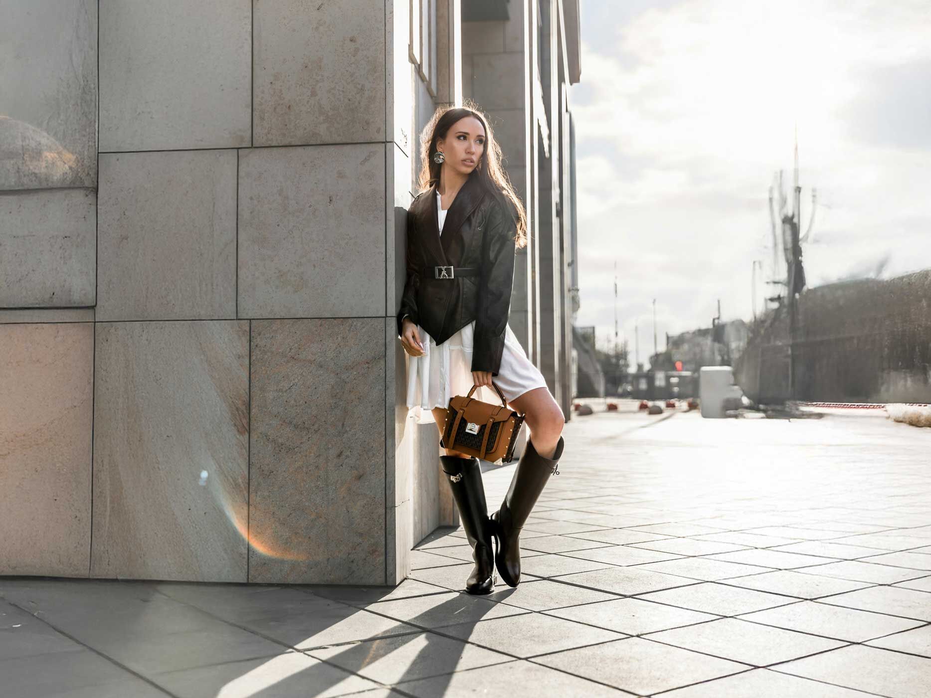 Eine Frau trägt ein knielanges weißes Kleid, dazu lange schwarze Stiefel, eine schwarze Lederjacke und eine braun-schwarze Lederjacke.