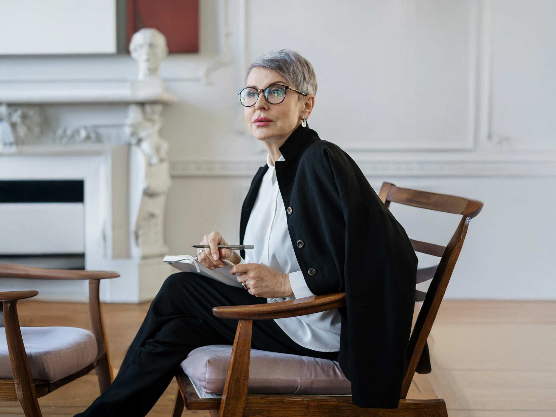 Eine Frau sitzt auf einem Stuhl, sie hat kurze graue Haare, trägt eine schwarze Brille, eine schwarze Hose, eine weiße lange Bluse und hat einen schwarzen Blazer über ihre Schultern gelegt, sie hält einen Stift und Papier in der Hand.