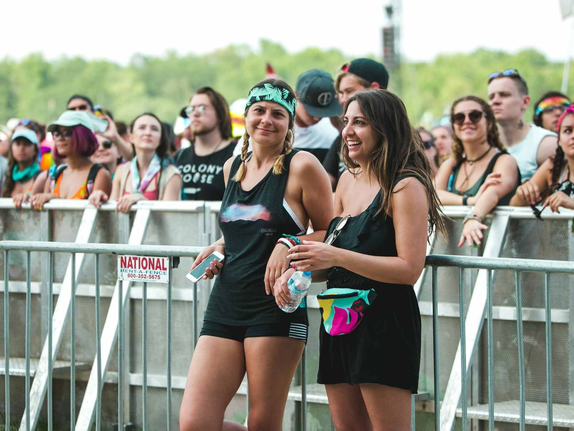 Zwei Frauen stehen in einer Menschenmenge vor den Absperrbarrieren, sie tragen ein schwarzes T-Shirt und eine schwarze Hose, bzw. einen schwarzen Jumpsuit. Sie haben eine Bauchtasche und Bandanas um.