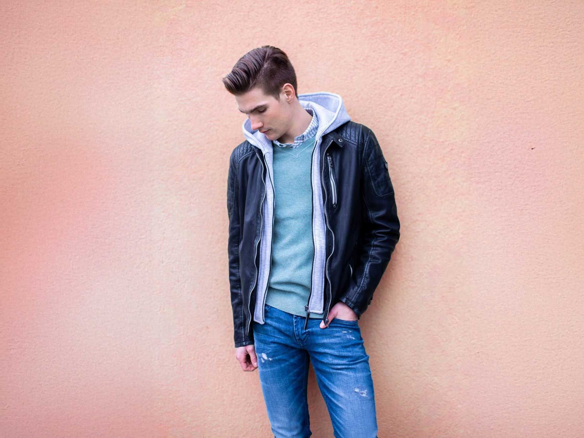 Ein junger Mann steht vor einer Hauswand, er trägt blaue Jeans, einen grünen Pullover, darüber eine graue Sweatjacke und eine schwarze Lederjacke.