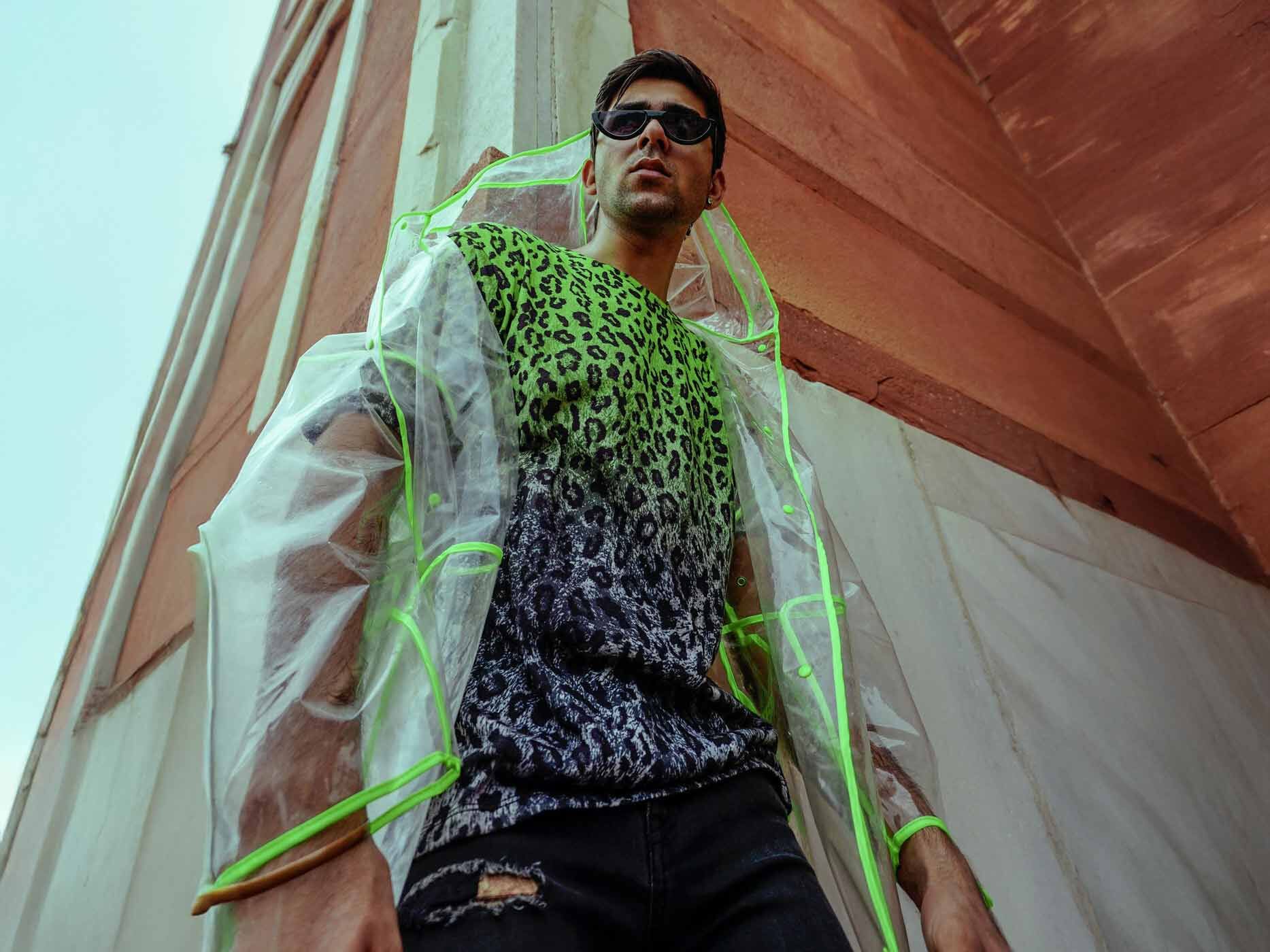 Ein Mann trägt eine schwarze Jeans, ein neon-grünes Leoprint-Oberteil und einen durchsichtigen, mit grünen Streifen verzierten Regenmantel darüber.