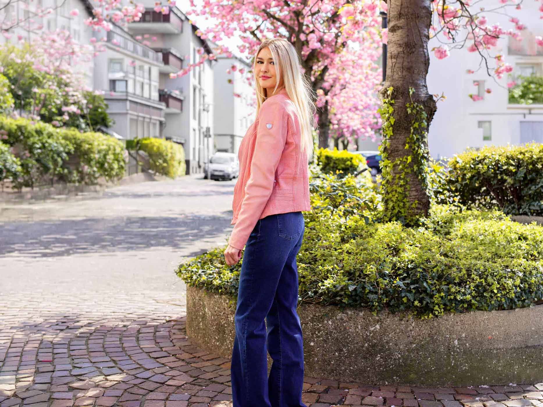 Eine junge Frau in rosa Lederjacke steht auf einem breiten Bürgersteig in der Stadt, im Hintergrund sind Häuser und rosa Kirschblüten