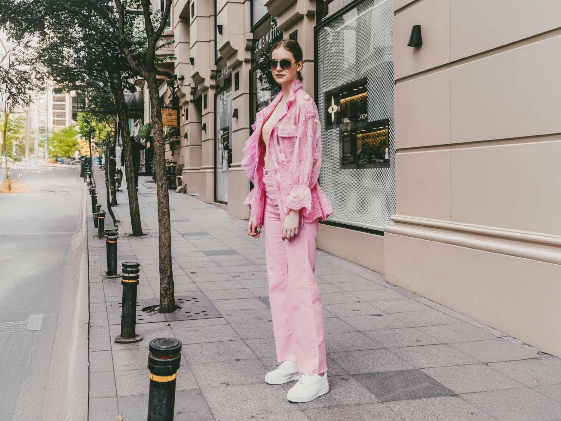 Eine Frau steht auf einem Gehweg, sie tr&auml;gt eine rosa Hose, wei&szlig;e Sneakers und eine rosa Jacke, dazu eine schwarze Sonnenbrille.