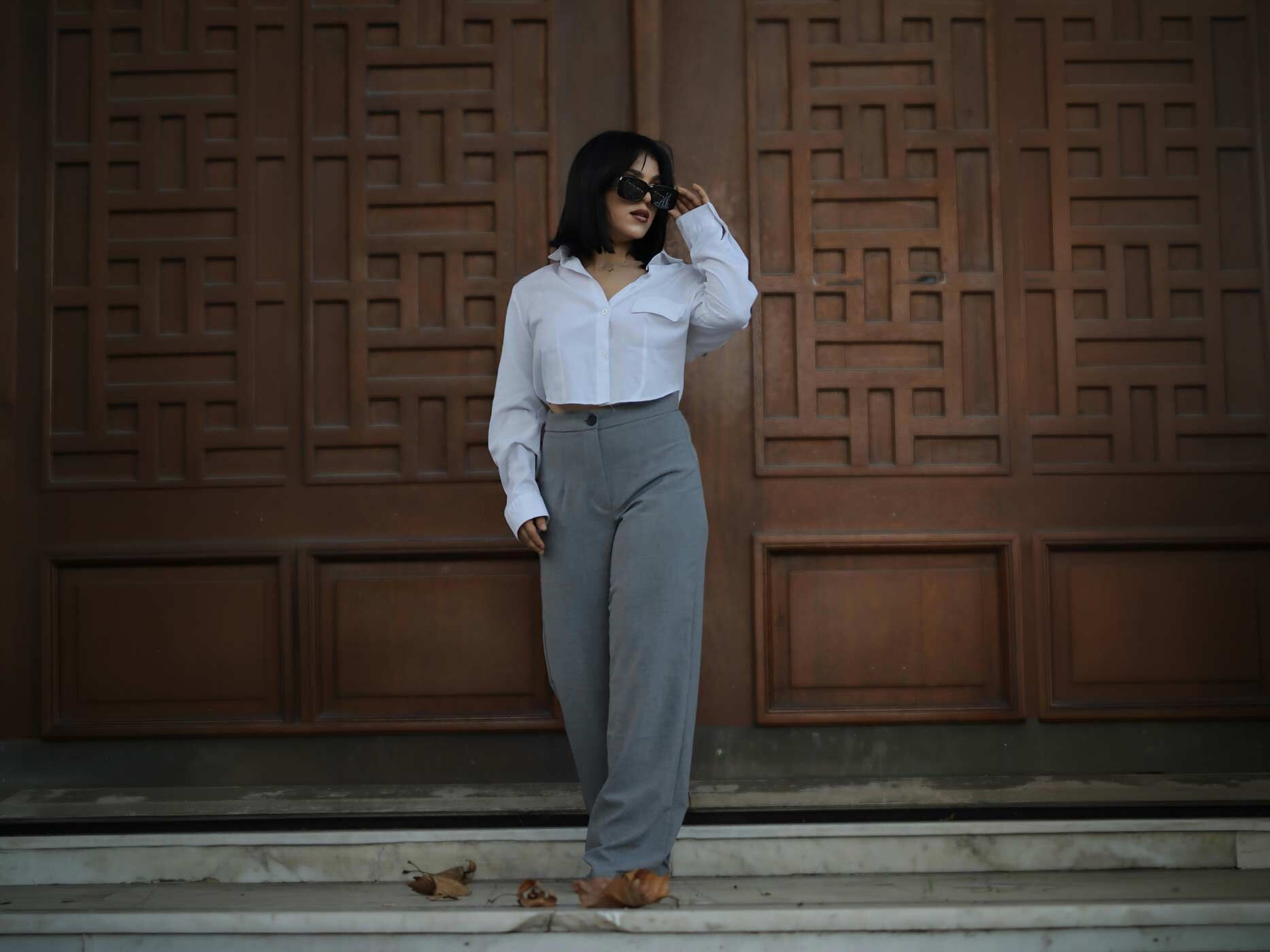 Eine Frau trägt eine graue Stoffhose und dazu eine kurze weiße Bluse.
